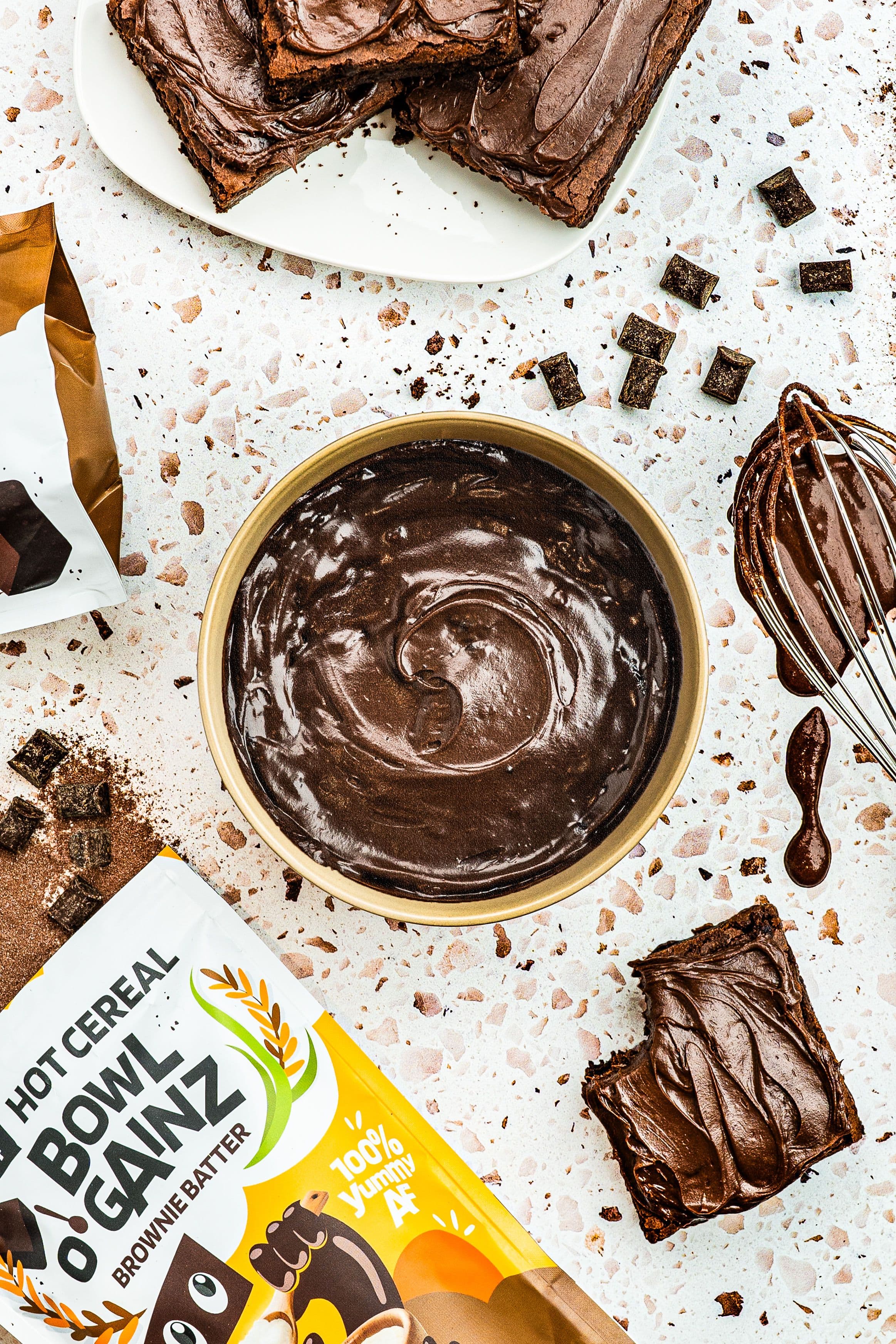 The Flavor Gang Brownie Batter Bowl O' Gainz cream of rice cereal cooked into a smooth, glossy chocolate batter in a gold bowl, surrounded by frosted brownies, chocolate chunks, and a batter-coated whisk.